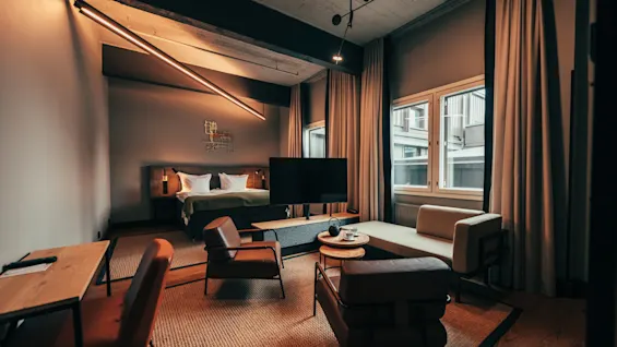 A stylish and modern hotel room with a comfortable bed, a seating area, a desk, and large windows, bathed in warm lighting.
