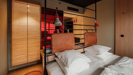 Bed with white pillows rests against a metal frame, featuring leather padding. Orange light fixtures hang nearby in a room with wooden blinds and warm-toned walls.