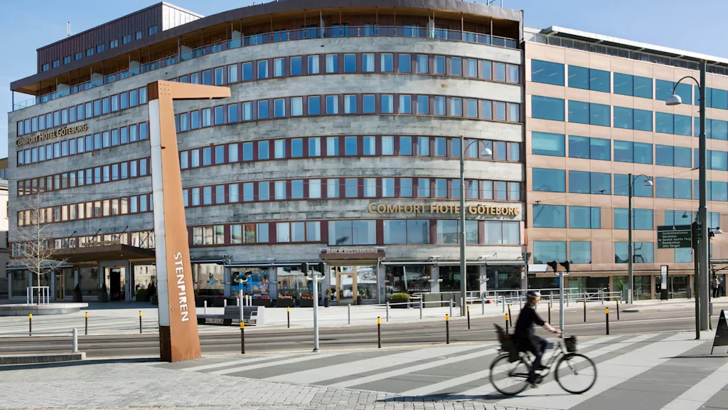 Polkupyöräilijä ajaa Comfort Hotel Göteborgin ohi, kulmikas rakennus heijastavine ikkunoineen aurinkoisena päivänä. Lähellä on puinen veistos, jossa lukee "STENPIREN".