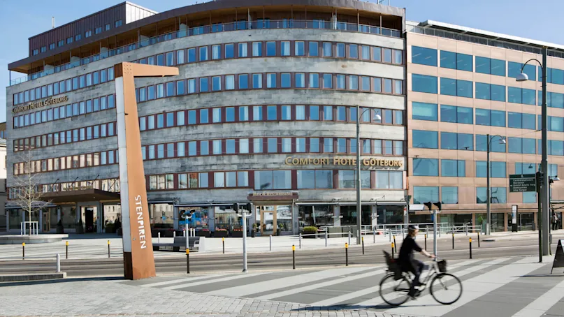 A cyclist rides past Comfort Hotel Göteborg, an angular building with reflective windows on a sunny day. Nearby is a wooden sculpture marked "STENPIREN".