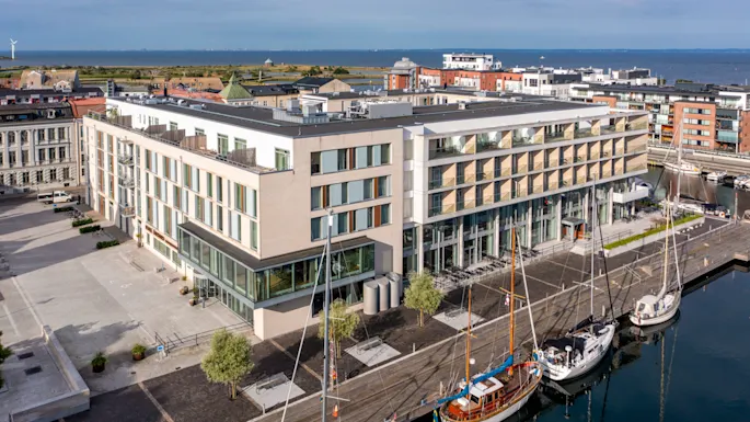 Moderni hotellirakennus lasijulkisivulla, josta on näkymä satamaan, jossa on useita veneitä, kaupunki ja meri taustalla.