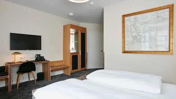 Et lyst og moderne hotelværelse med to komfortable senge, et skrivebord med TV og et dekorativt indrammet kort.