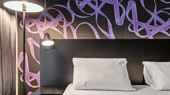 A bed with white linens against a dark headboard, illuminated by bedside lamps, with abstract purple art on the wall.