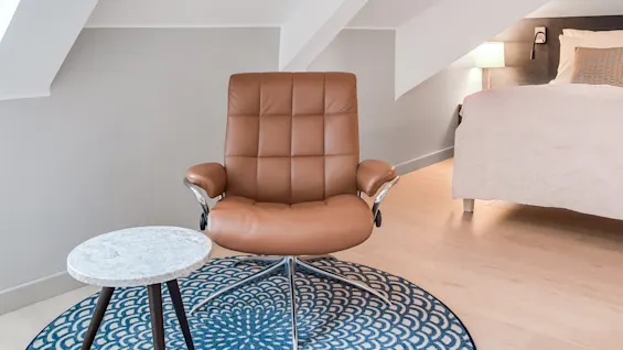 En brun lænestol står på et blåt mønstret tæppe med et lille sidebord. I baggrunden, en seng med en hvid lampe i et rum med skrå loft.