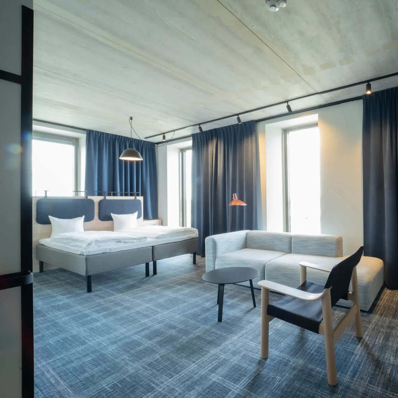Moderni hotellihuoneessa on sänky, sohva ja pieni pöytä hyvin valaistussa tilassa, jossa on siniset verhot ja matto.