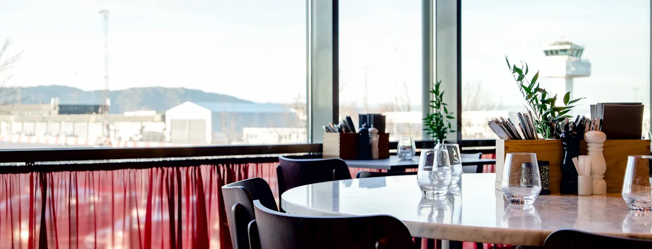 En restaurant med borde og stole, med et stort vindue, der vender ud mod et lufthavnstårn.
