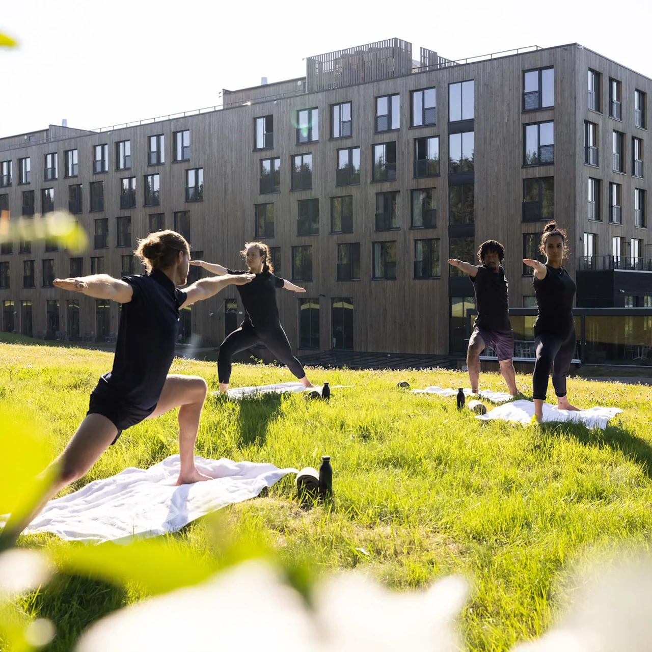 Personer tränar tillsammans på en gräsmatta framför moderna byggnader, utför yoga eller liknande sträckövningar i solsken på Lily Country Club i norska Klöfta.