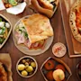 Ett urval av italienska rätter ligger utbrett på ett träbord, inklusive pizza, smörgås, sallad, oliver och arancini, vilket skapar en livfull och aptitretande matsituation typisk för italiensk mat.