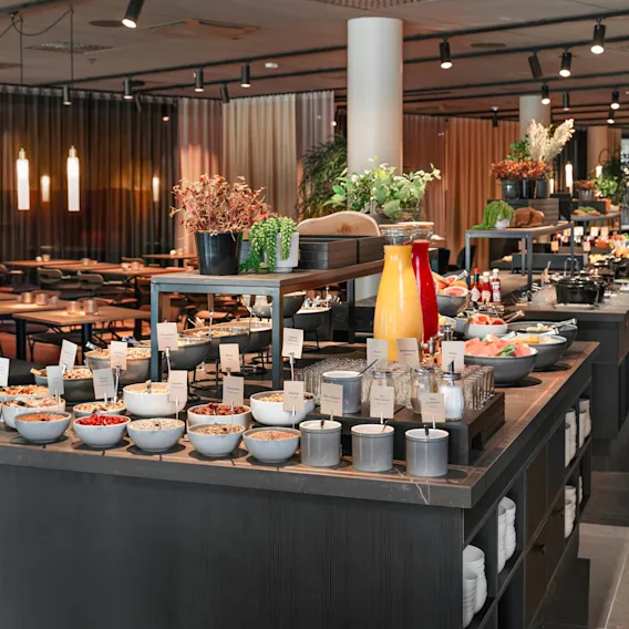 Buffet table displaying assorted breakfast items like cereals, juices, fruits, and condiments in a well-lit restaurant with plants and modern seating arrangements.