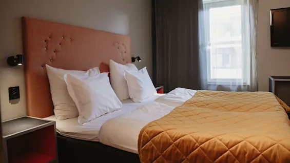 En säng med vita kuddar och ett orange, vadderat täcke står i ett hotellrum. Den har ett stoppat huvudgavel, sängbord, vägglampor, ett fönster och en TV.