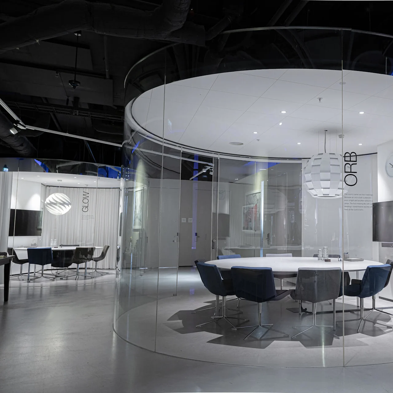 A round conference table with chairs sits in a glass-walled room. "ORB" and "GLOW" are on walls. The room is modern with a clock and television.