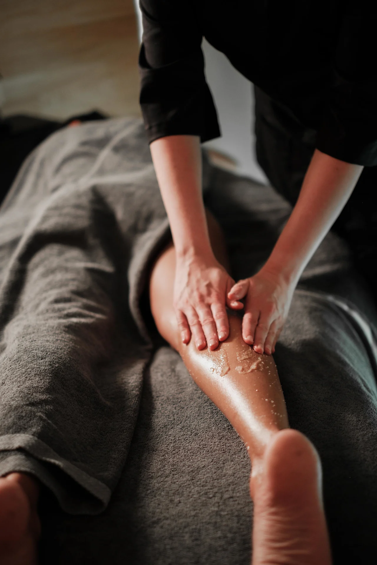 En massageterapeuts händer gnider in ett exfolierande skrubb på en klients ben, som ligger täckt med en grå handduk i en svagt upplyst miljö.