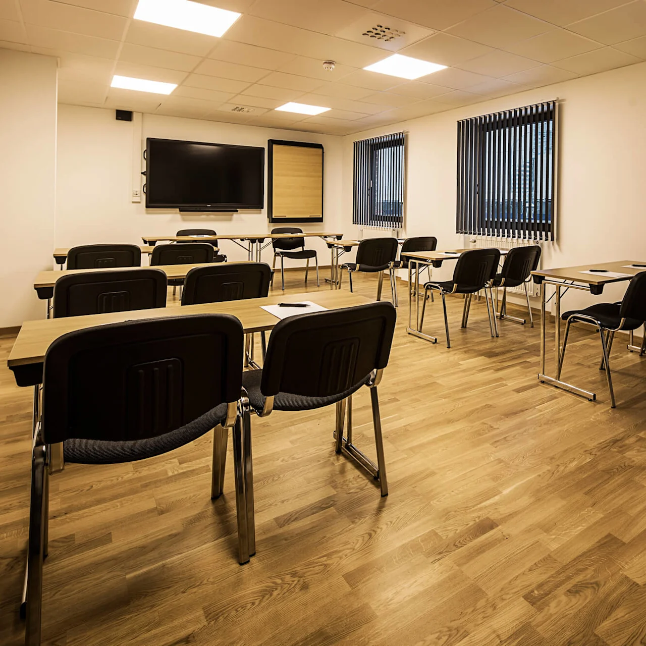 Conference room with desks and chairs facing a large screen, white walls and windows with vertical blinds.