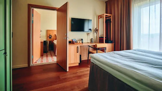 Et koselig hotellrom med en komfortabel seng, et skrivebord med TV og en åpen dør som viser et tilstøtende rom.