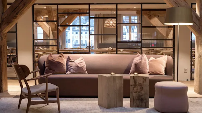 Sofa with cushions sits on a rug, flanked by wooden tables and a lamp. Background features large windows and wooden beams, with visible dining area.