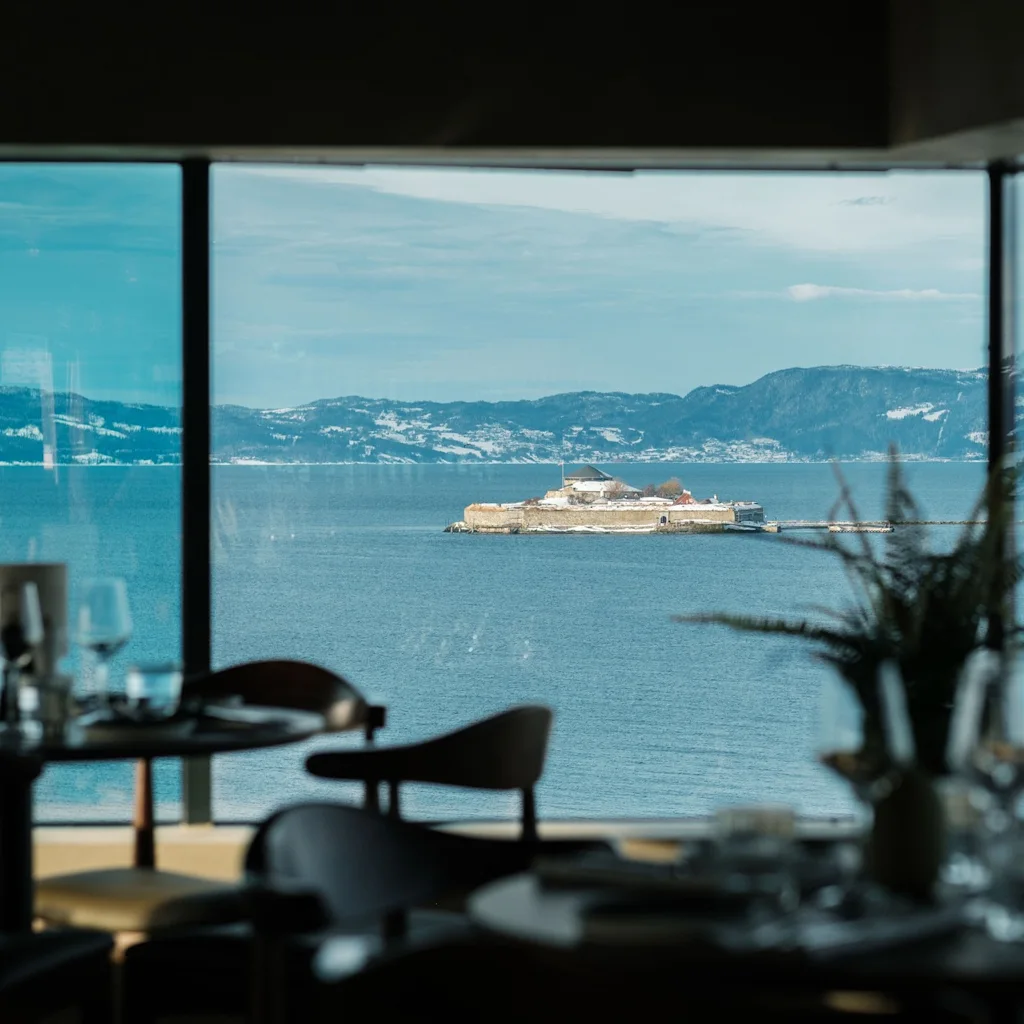 Restaurantinteriør med en fantastisk vindusutsikt over en rolig innsjø, en liten øyfestning og majestetiske snødekte fjell.