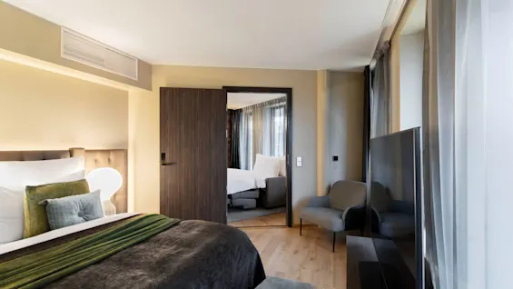 A neatly arranged bedroom features a plush bed with cushions, facing a large flat-screen TV. A door opens to reveal another bedroom with similar decor, under soft lighting.