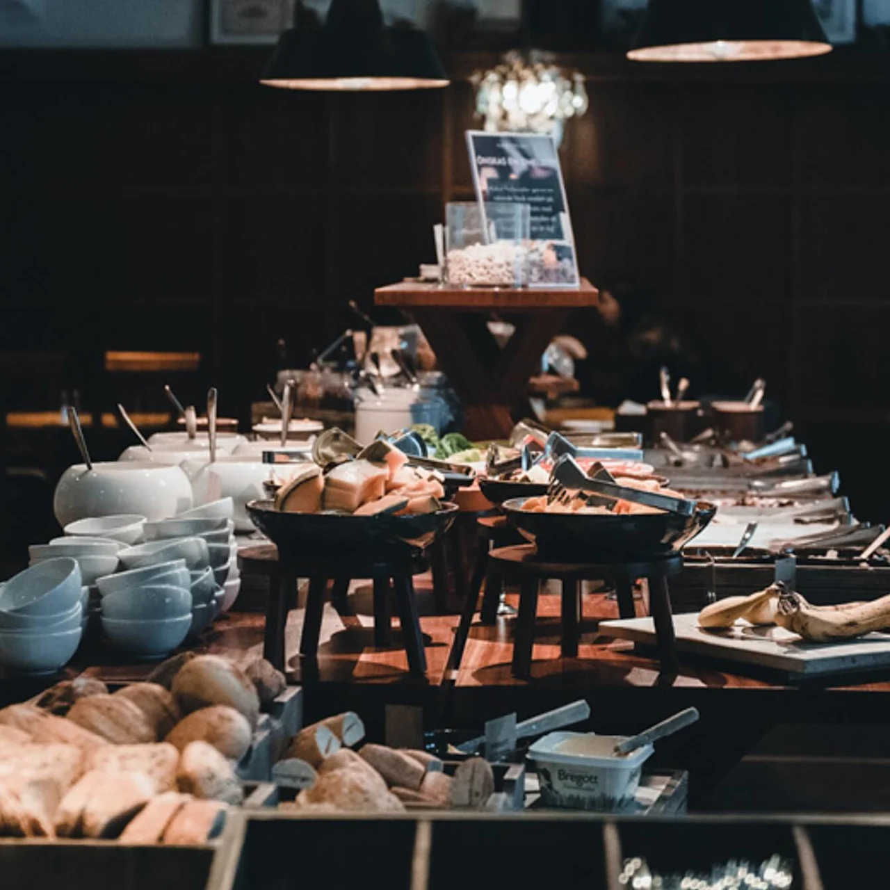 Et buffetbord viser forskellige brød, frugter og pålæg. Assorterede skåle og tallerkener er arrangeret i nærheden, beliggende i en hyggelig spisestue med varm belysning.