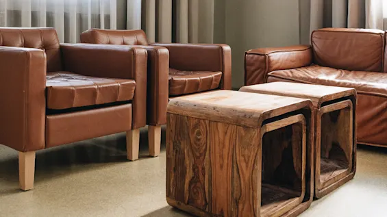 Brown leather armchairs and a sofa are arranged around wooden cube tables in a room with beige curtains and large windows. A ceiling lamp hangs overhead.