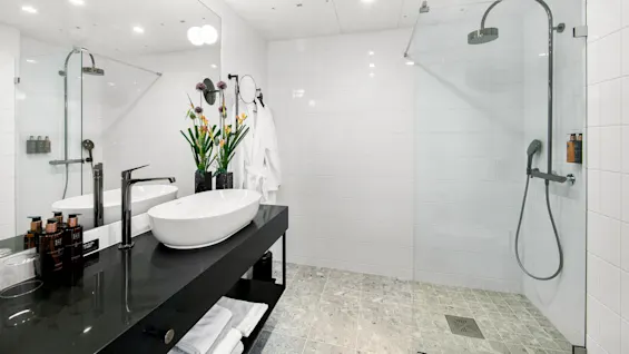 Sleek bathroom with a modern sink on a black countertop adorned with flowers. A glass shower enclosure is to the right, with white tiled walls and floor. Robes hang nearby.