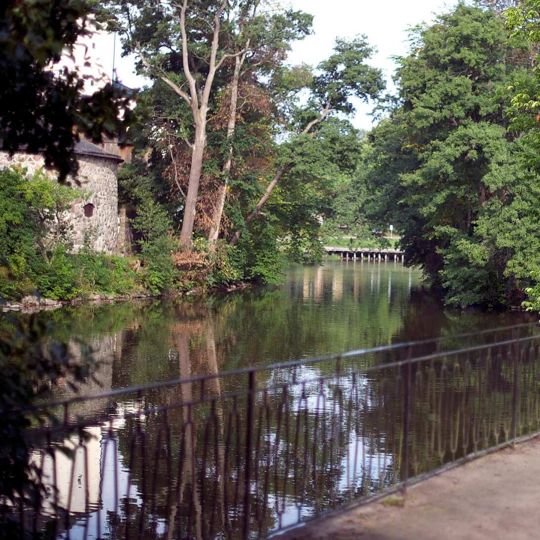 A serene pathway beside a calm river, with a lush green backdrop and a large flower pot on the railing.