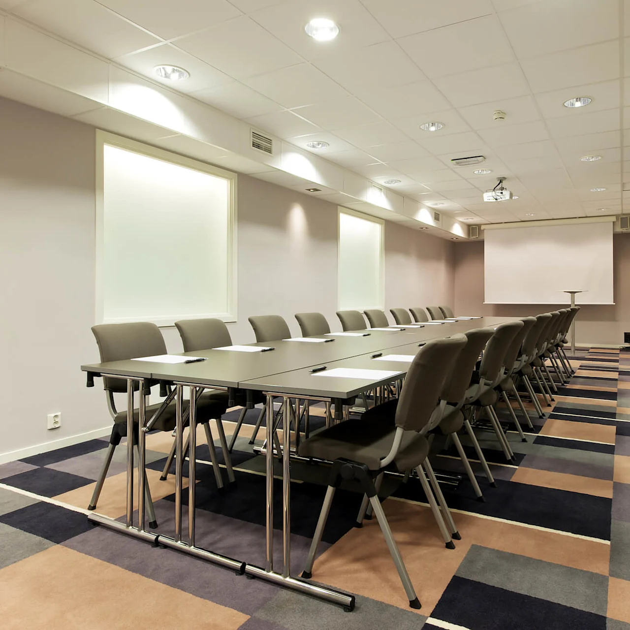 Et langt konferansebord med stoler, papir og penner, vendt mot et projeksjonslerret i et teppebelagt møterom med belysning.