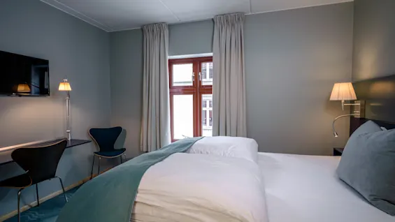 En seng med hvidt sengetøj og et blåt tæppe står i et moderne rum. Et skrivebord, stole og et vægmonteret TV er i nærheden; lys strømmer gennem et gardineret vindue.