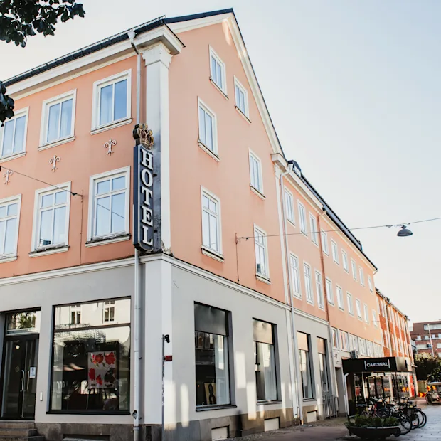 Persikanvärinen hotellirakennus, jonka kulmassa on näkyvä pystysuora HOTEL-kyltti, sijaitsee aurinkoisella kadulla muiden rakennusten ja pysäköityjen polkupyörien kanssa.