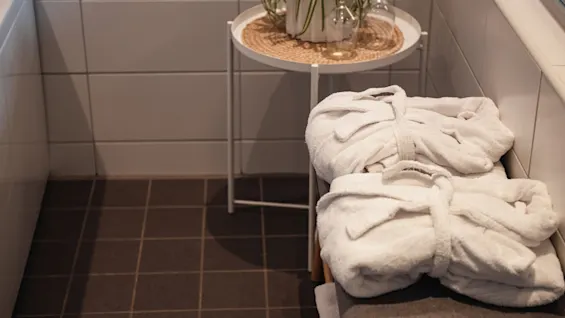 Two plush white bathrobes await on a bench in a tiled bathroom, next to a small table with a plant and glasses, suggesting relaxation.