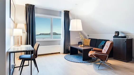 Hyggelig stue med en brun læderstol vendt mod et vindue, der viser en naturskøn vandudsigt, omgivet af en sofa, sofabord, skrivebord og varm belysning.