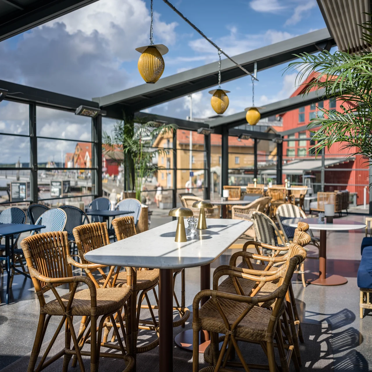 Rottingsstolar omger ett vitt marmorbord under gula hänglampor i ett glasomslutet café med utsikt över en hamn och färgglada byggnader.