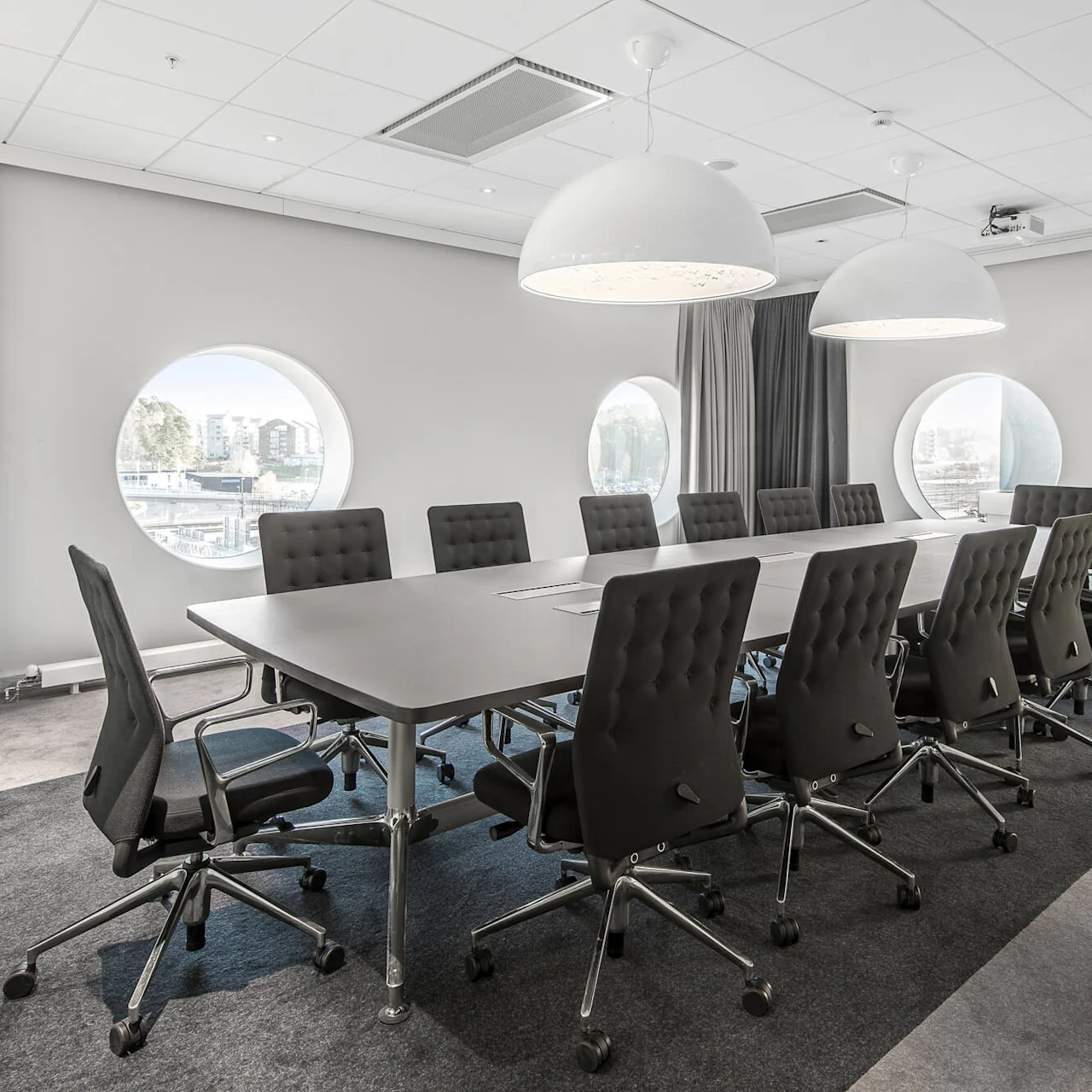 Et langt, rektangulært konferencetable er omgivet af sorte drejestole, placeret i et moderne rum med cirkulære vinduer og kuppellamper, der giver lys.