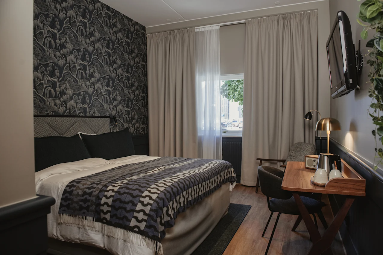 Ett mysigt hotellrum med en mönstrad accentvägg, en bekväm säng med en dekorativ pläd, ett litet skrivbord och ett fönster.