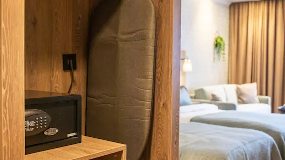 Strykningbräda och kassaskåp i en trägarderob; en mini-kyl finns i närheten. I bakgrunden syns ett mysigt hotellrum med bäddad säng och blå sängkläder samt varm belysning.