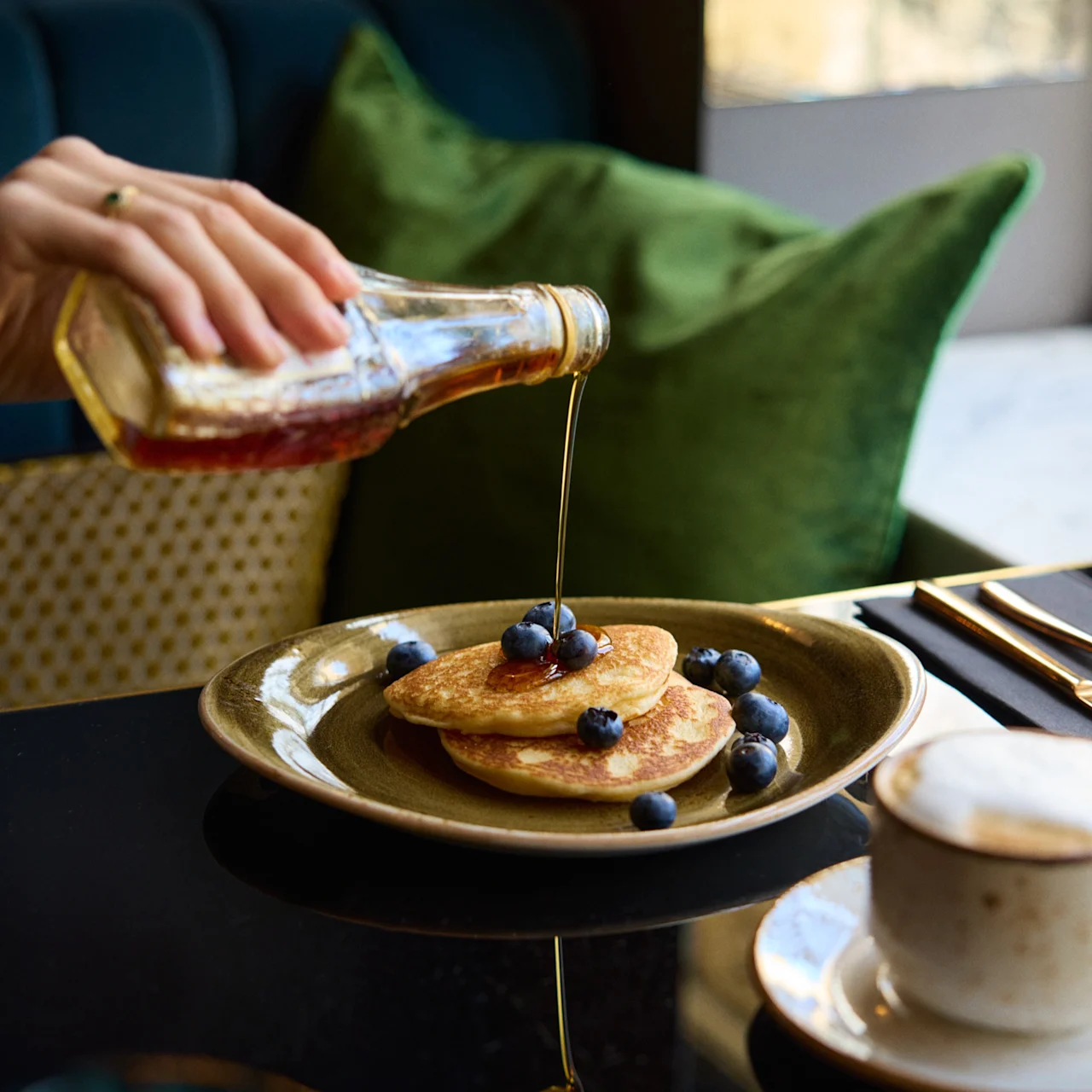 Pancakes with blueberries being drizzled with syrup by a hand, on a table with a coffee cup, green cushions, and soft lighting.