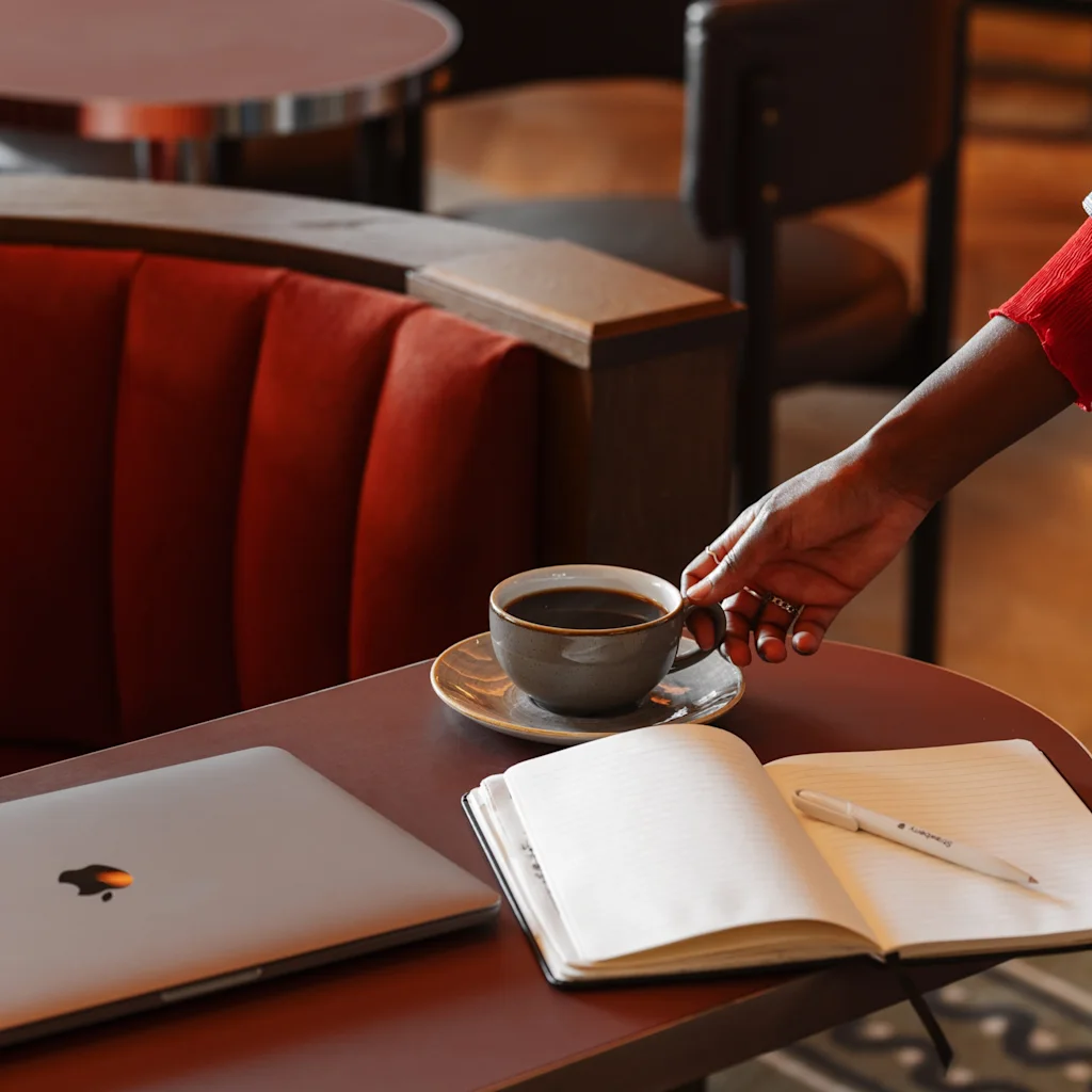 En hånd strekker seg etter en kaffekopp på et bord med en bærbar datamaskin og åpen notatbok, i et komfortabelt salongområde.