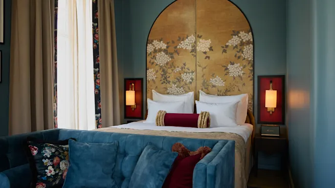 A bed with floral-patterned headboard sits in a cozy room. Two bedside lamps provide warm light. A blue sofa with decorative pillows faces the bed, creating a relaxing atmosphere.