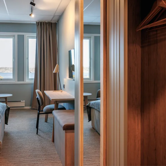 Et koselig hotellrom har en seng, skrivebord og stol. Store vinduer med gardiner viser en havutsikt. Et tre-skap med kleshengere er delvis synlig.