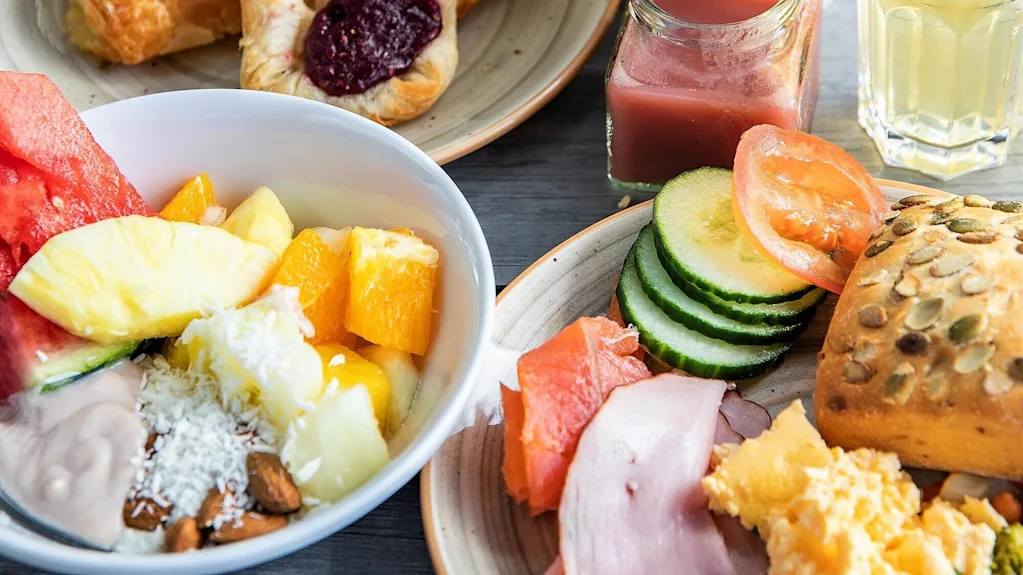 A diverse breakfast spread features pastries, fruit salad, meats, eggs, and bread on plates; accompanied by coffee, juice, and a vase of white flowers on a wooden table.
