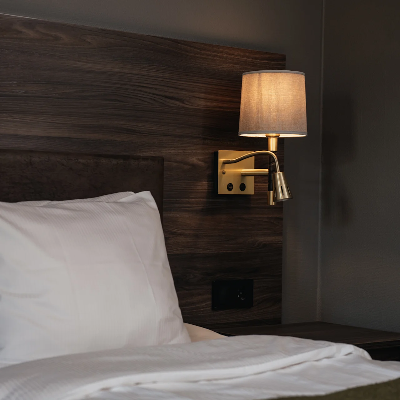 Bed with white pillow and bedding, situated beneath a wooden headboard. A wall-mounted lamp with a beige shade illuminates the setting, creating a cozy bedroom atmosphere.