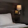 Bed with white pillow and bedding, situated beneath a wooden headboard. A wall-mounted lamp with a beige shade illuminates the setting, creating a cozy bedroom atmosphere.