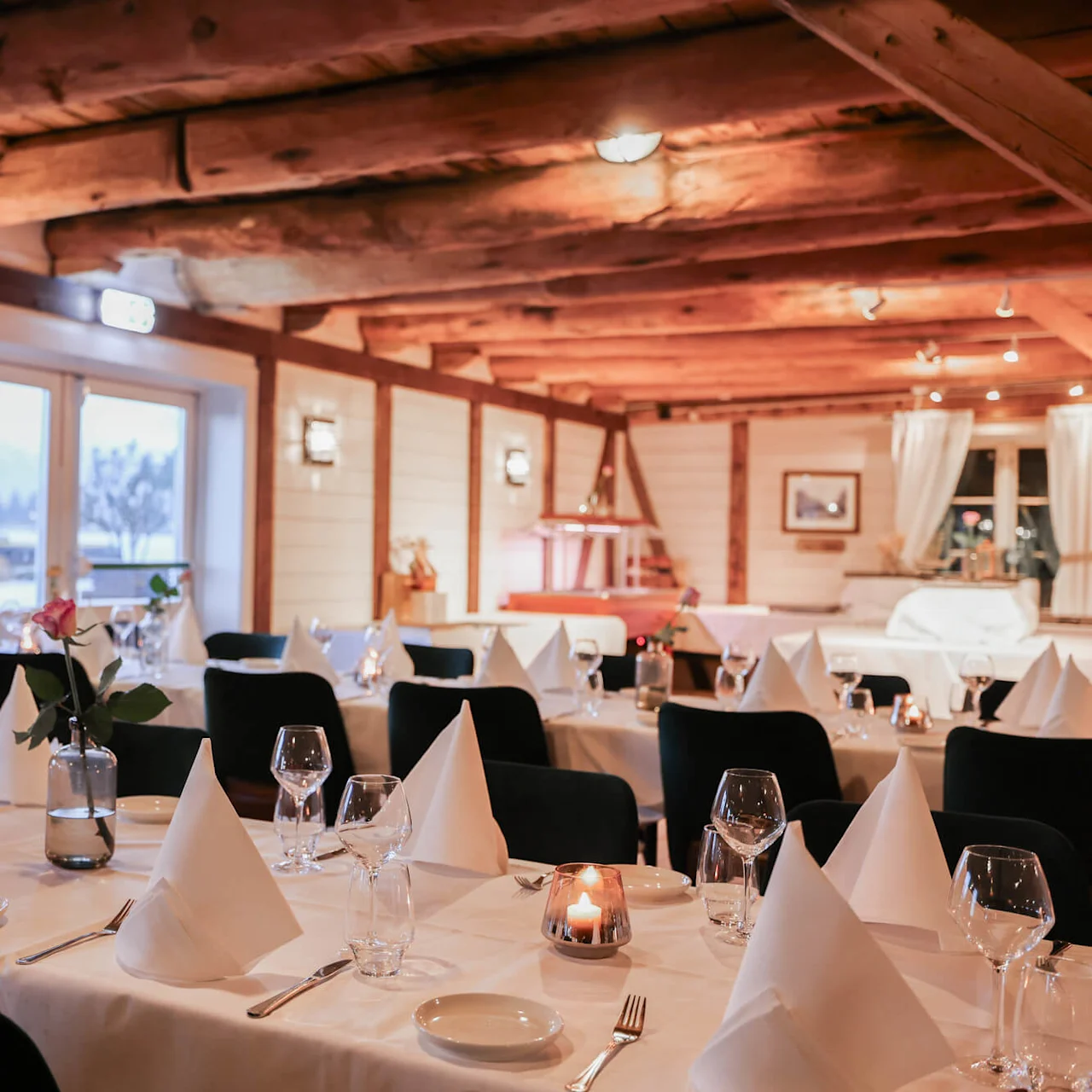 Elegant dining tables are set with white cloths, neatly folded napkins, and glassware in a cozy, rustic restaurant with exposed wooden beams and soft lighting.