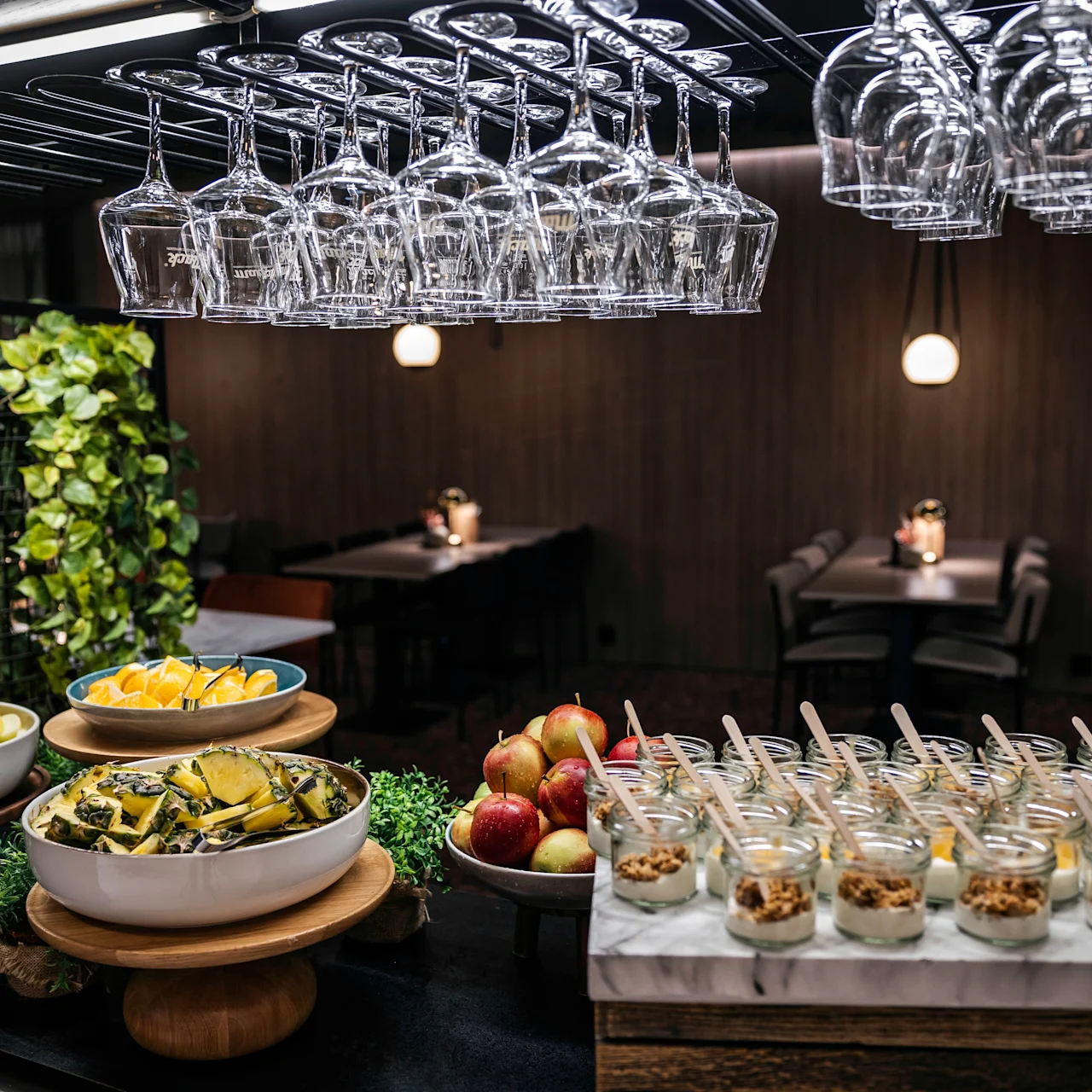 Glasses hang above a buffet table with fruit bowls and yogurt jars, surrounded by indoor plants. Dimly lit dining tables are visible in the background.