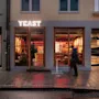 A vibrant restaurant Yeast at dusk with people dining inside, and a person walking on the wet sidewalk outside.