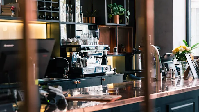 A coffee shop counter displays a modern espresso machine, surrounded by mugs and glassware. Nearby, plants and flowers decorate the countertop, creating a welcoming atmosphere.