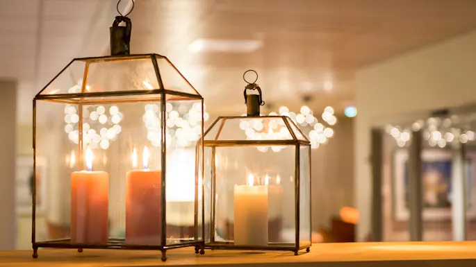 Two glass lanterns with lit candles inside, creating a warm ambiance against a softly lit bokeh background in an indoor setting.