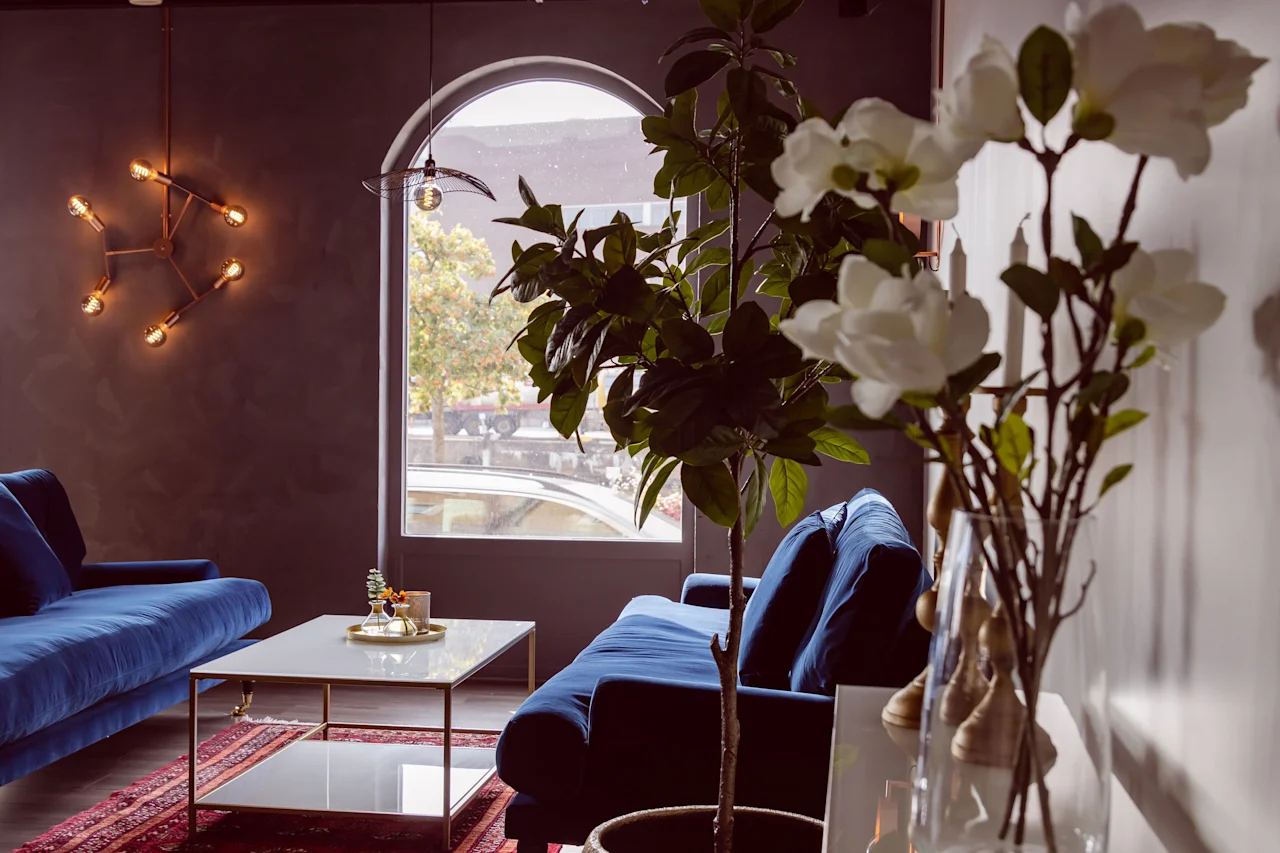 Mysig hotellounge med blå sammetssoffor, varm belysning, gröna växter och ett stort bågformat fönster.