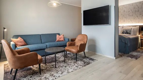En blå sofa med orange puder står overfor to brune lænestole og et rundt bord på et mønstret tæppe. Et TV er monteret på væggen; et soveværelse er delvist synligt.