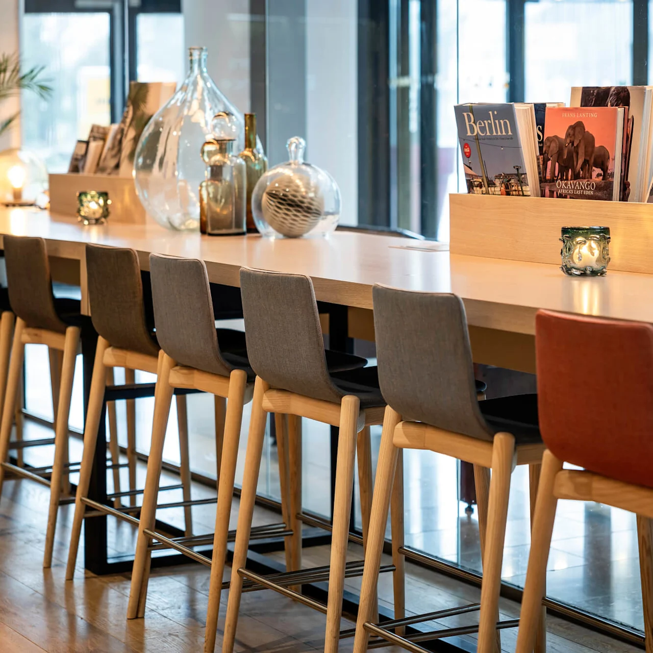 Langt trebord med høye stoler, med dekorative glassflasker og magasiner. Titler inkluderer "Berlin" og "Okavango". Ligger i et moderne, lyst innendørsområde med store vinduer.