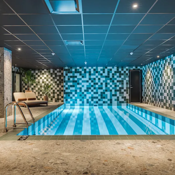 En inbjudande inomhuspool med blå och vitrandiga plattor, omgiven av rutiga väggar och solstolar.
